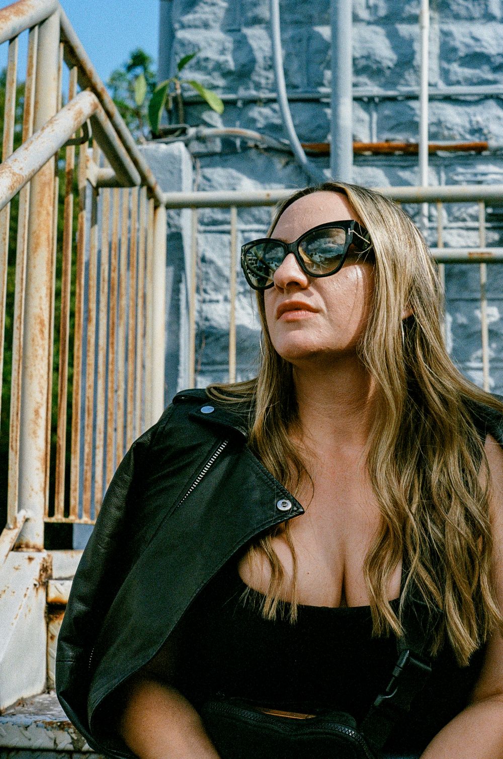 A person with long blonde hair wearing sunglasses and a black leather jacket poses against a stone wall outdoors.