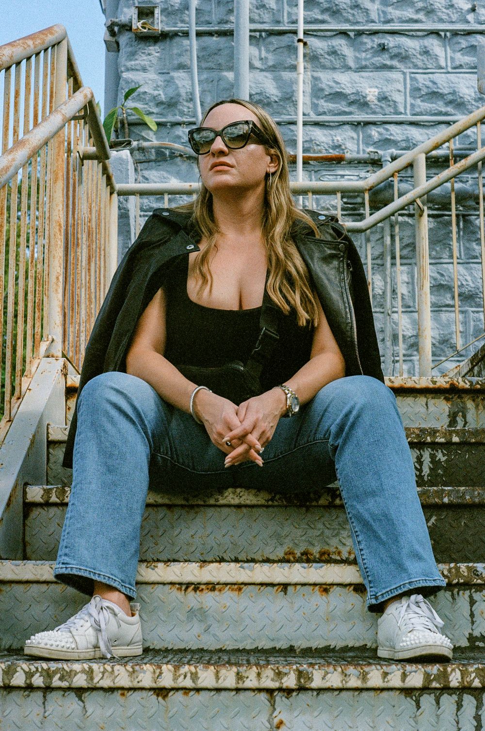 Stylish woman in leather jacket and sunglasses sits casually on concrete steps against stone building exterior.