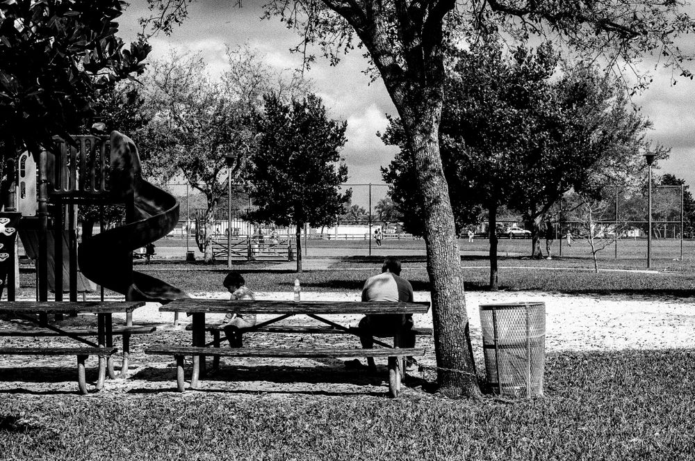Black and white photo of a playground with picnic tables and trees in a public park setting.