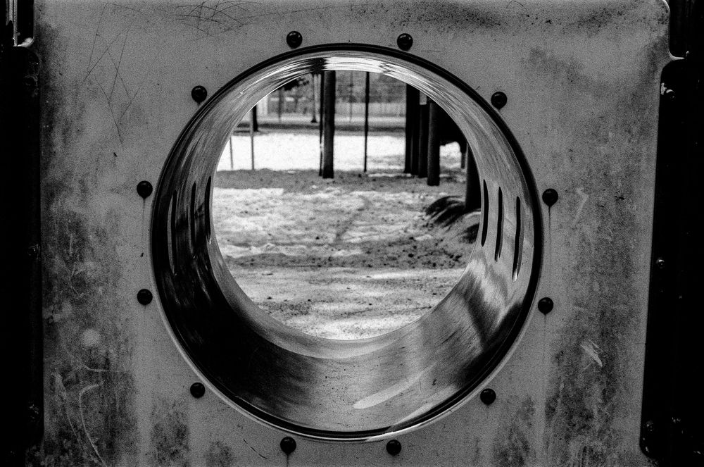 A black and white view through a circular porthole or tunnel opening shows a playground slide in the distance.