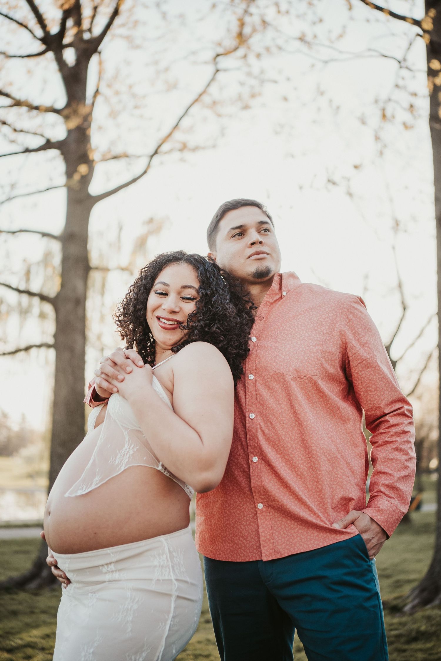 A couple poses together outdoors during a maternity photoshoot, with the pregnant woman in white and partner in coral shirt.