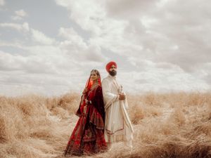 A couple wearing traditional South Asian wedding attire stands in a golden wheat field under cloudy skies.