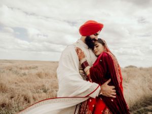 A couple in traditional Punjabi wedding attire embrace in a golden field under cloudy skies.