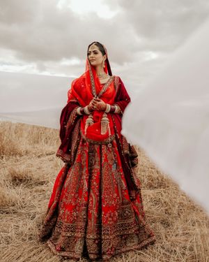 A bride in a traditional red and gold embroidered lehenga poses dramatically against a cloudy sky in a field.
