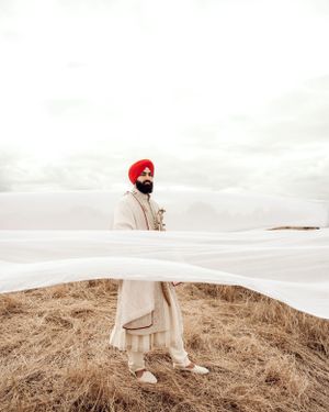 A person in a red turban and beige coat stands in a field while white fabric billows in the wind on a cloudy day.
