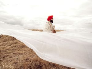 A figure wearing a red turban stands amid flowing white fabric billowing in the wind against a moody sky and dry landscape.