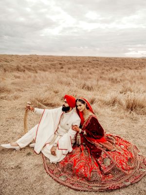 Traditional Sikh wedding couple sitting in a field wearing ornate red and white cultural attire during their photoshoot.