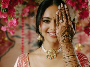 Beautiful bridal mehndi design on hand shown against backdrop of pink flowers and traditional Indian wedding attire and jewelry.