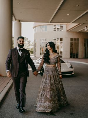 Couple in traditional Indian wedding attire walking hand in hand near a luxury car under covered walkway.