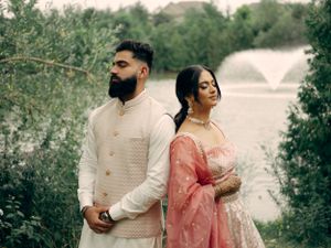 A couple in traditional South Asian attire sharing a romantic moment by a lake with green foliage in the background.