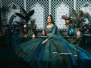 A woman in an ornate teal Victorian-style ball gown sits elegantly amidst decorative floral arrangements and lattice backdrop.