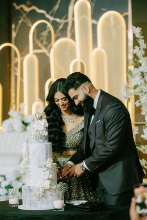 A couple cuts their elegant white wedding cake together in front of illuminated architectural arches.