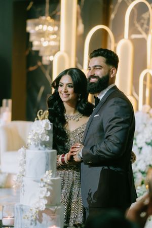 Couple in elegant Indian wedding attire stand together at reception with illuminated backdrop.