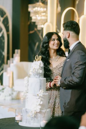 A couple stands beside an elegant white wedding cake decorated with floral details.