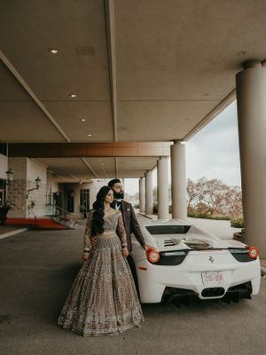 Newlyweds in traditional and modern fusion attire pose with white sports car under covered entrance.