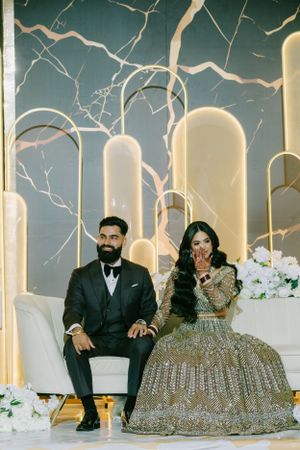 A couple sits together in formal attire against a backdrop of illuminated archways and decorative branches.