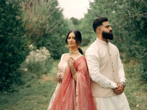 A romantic couple in traditional South Asian wedding attire pose together outdoors among trees.
