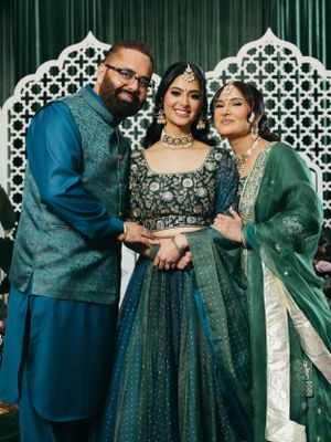 Three people wearing traditional Indian wedding attire in blue and green colors pose together against an ornate backdrop.