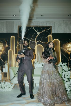 Two people posing at an elegant wedding reception with a sparkly gold backdrop and smoke effects in view.