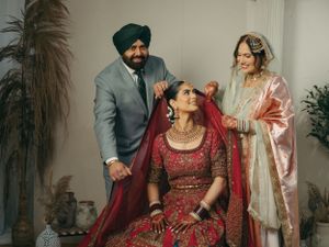 Traditional Indian wedding preparation scene with family members adjusting a bride's ornate red lehenga and jewelry.