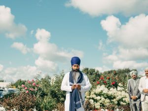 Someone wearing traditional Sikh attire stands in a garden with blooming flowers under a bright blue sky with white clouds.
