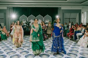 People dancing in traditional South Asian attire at a wedding celebration with decorative backdrop and audience.