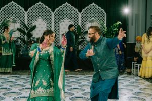 A couple in matching teal outfits dance energetically at an indoor celebration with decorative arches in the background.