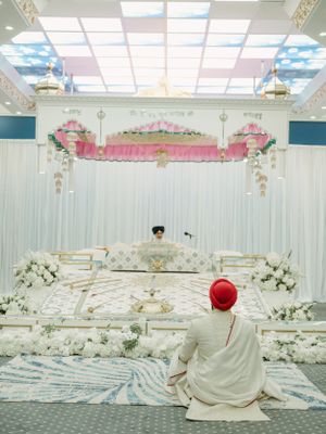 A Sikh wedding ceremony setup with white and pink decor, skylight ceiling, and someone seated in white attire.
