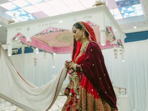 A traditional Indian bride wearing an ornate red and gold lehenga with embroidery details stands in an elegant wedding venue setup.