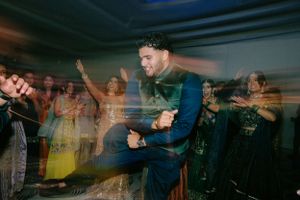 A performer energetically jumps on stage while blurred crowd dances in the background during a concert.