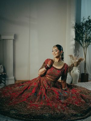 A bride in a luxurious red and gold embellished lehenga poses gracefully against a light backdrop with classical pillars.