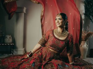 Traditional Indian bride in luxurious deep red lehenga poses elegantly against a red backdrop with white columns.