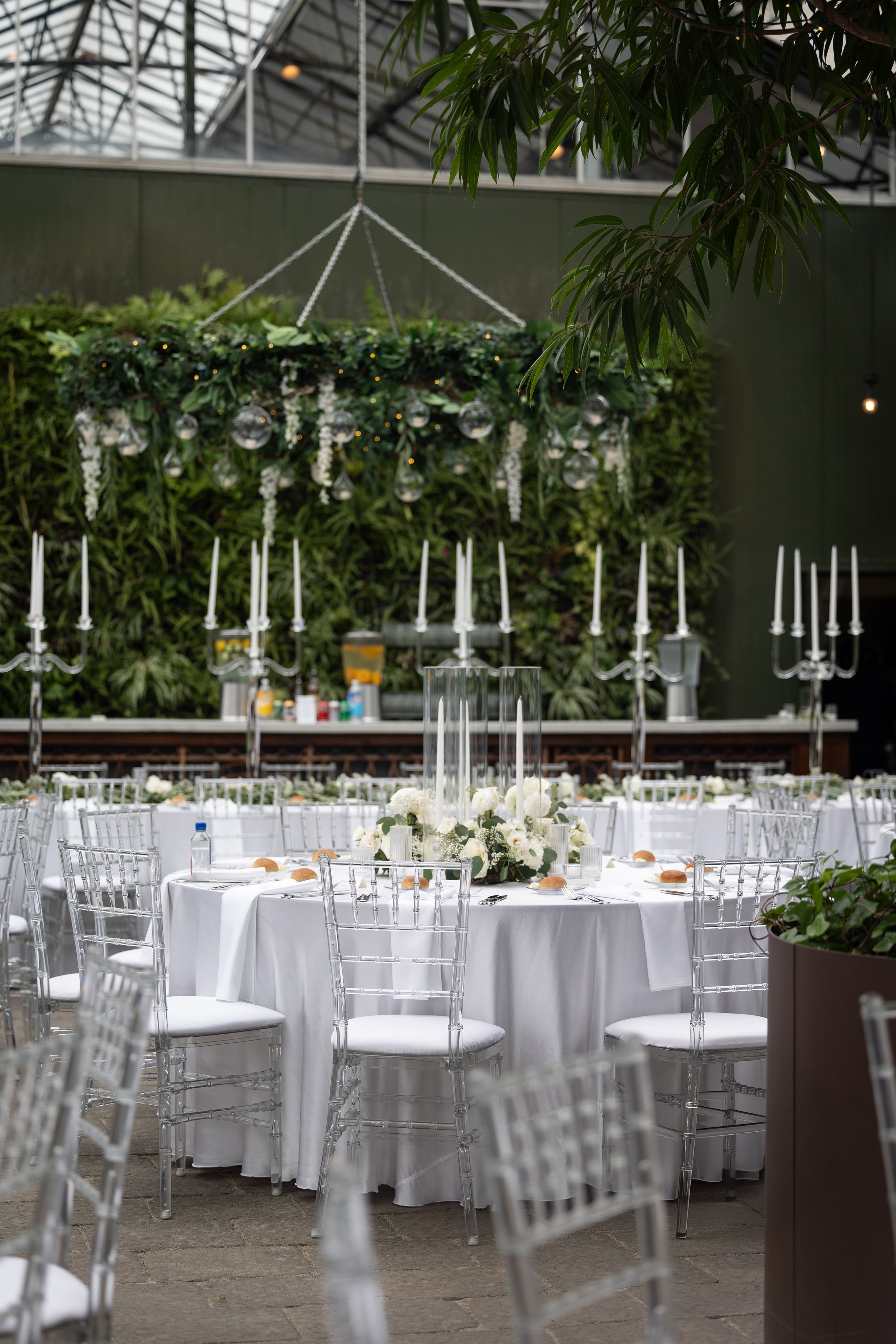 Elegant wedding reception setup with white tablecloths, silver chairs, and hanging floral arrangements in a greenhouse venue.