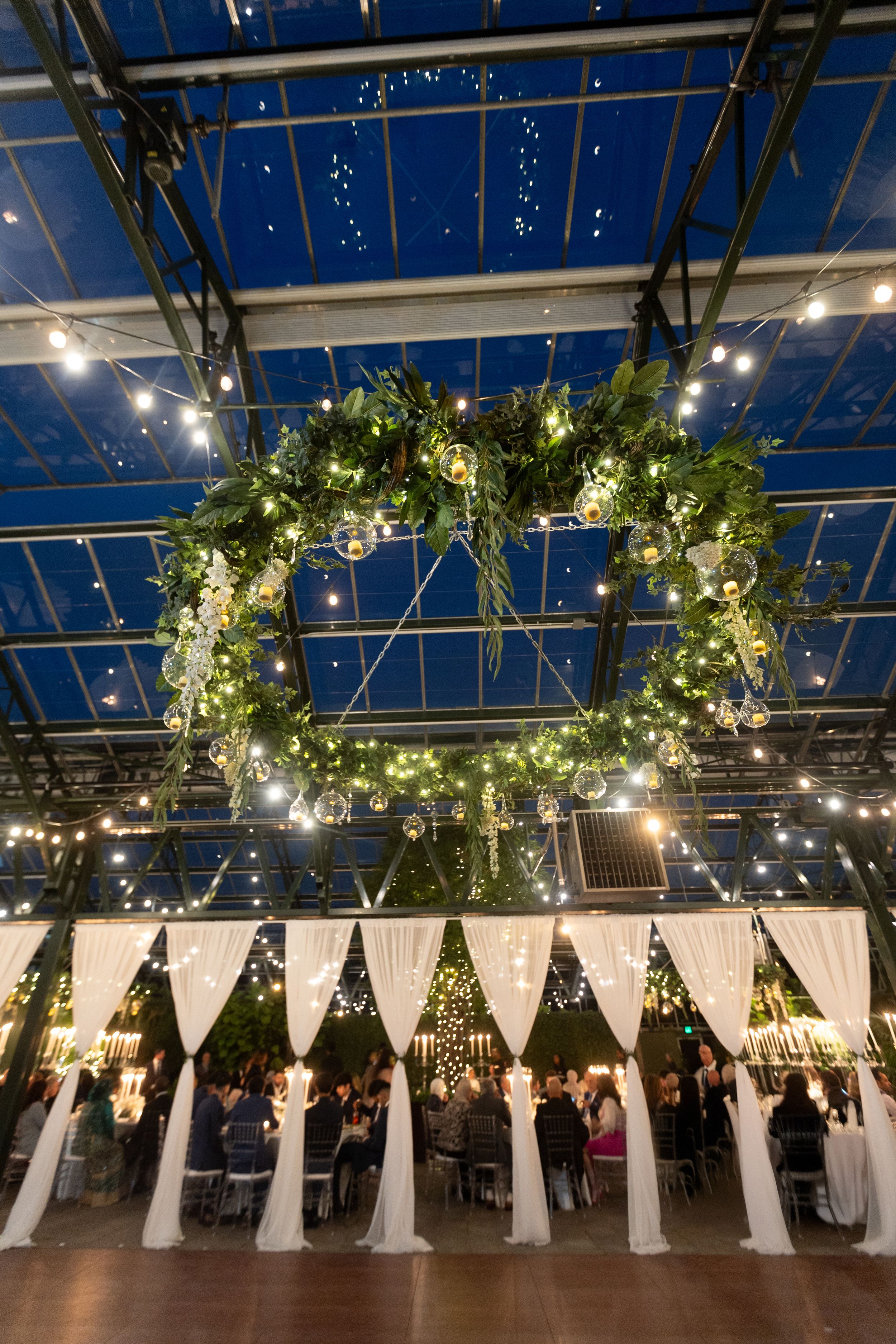 A large illuminated floral wreath hangs from a glass ceiling above white draped partitions at an elegant wedding reception.