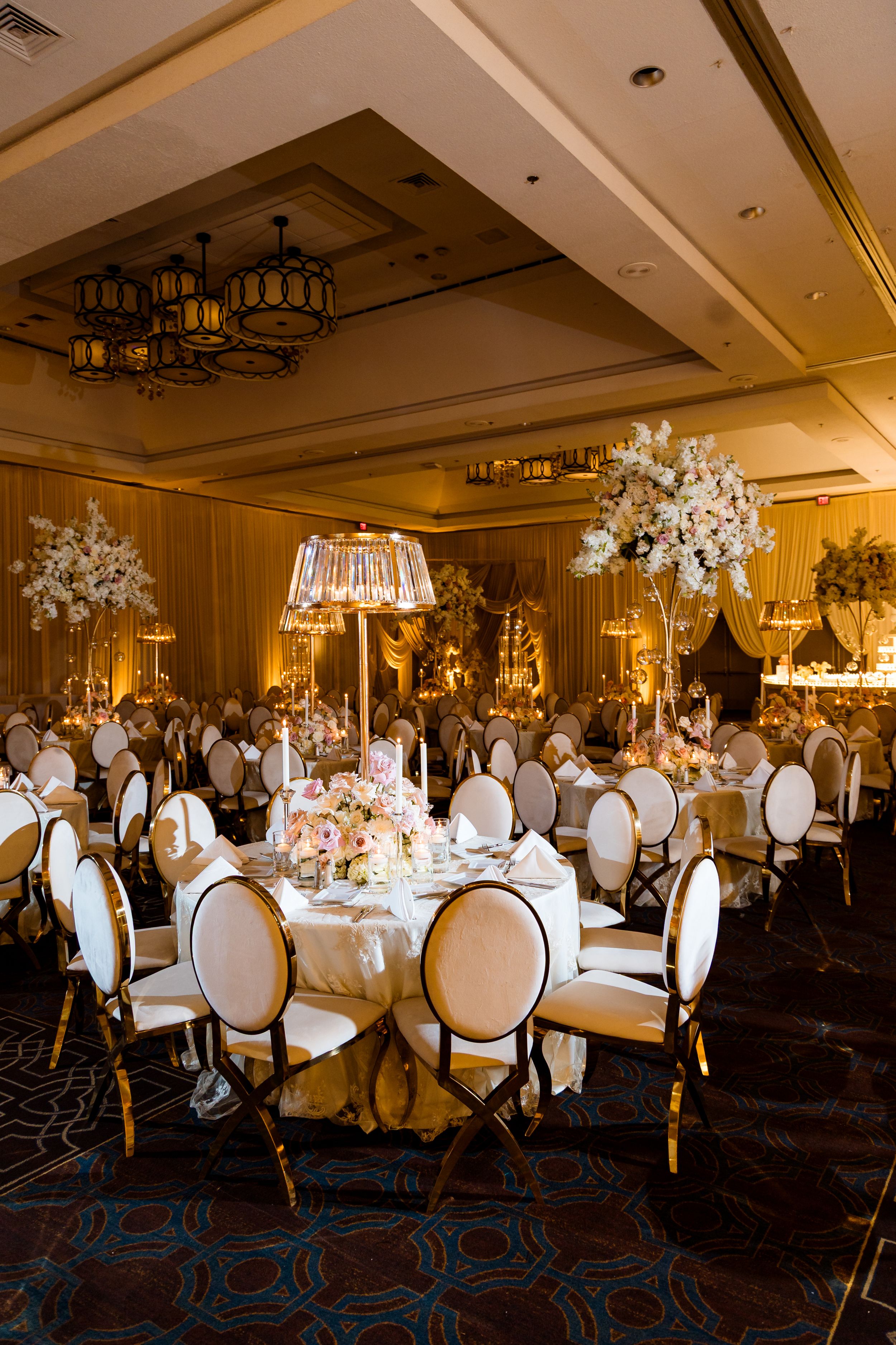 Elegant ballroom wedding reception setup with white chairs, crystal chandeliers, and floral centerpieces on round tables.