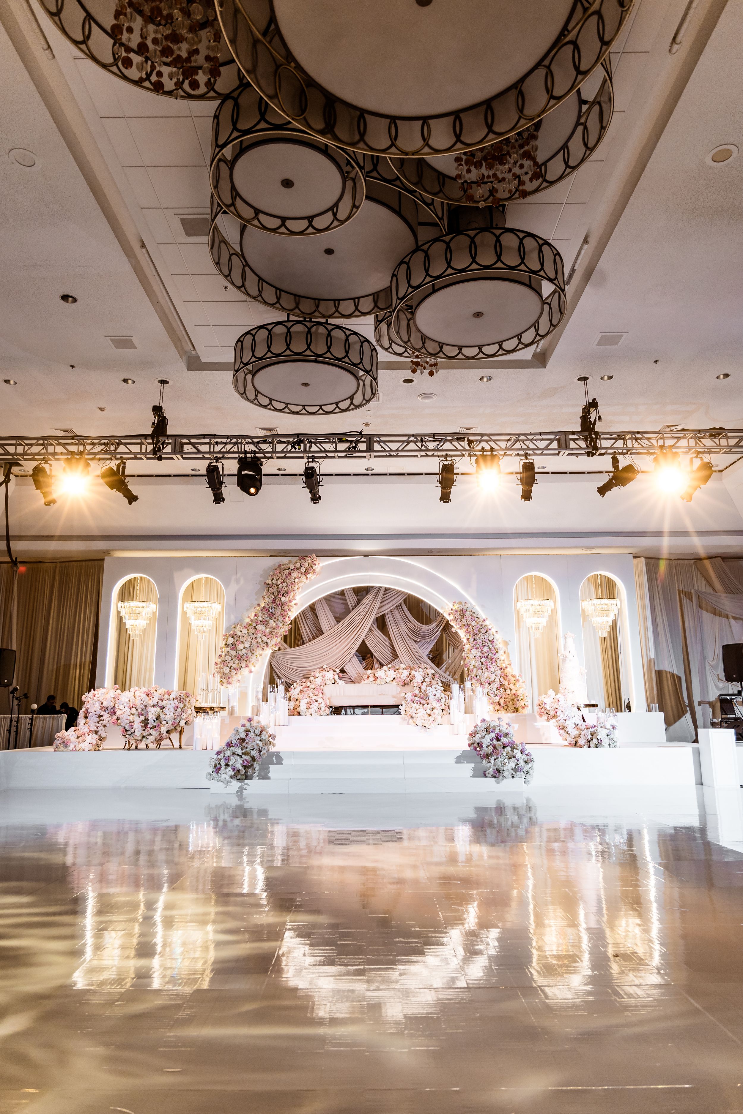 Elegant ballroom with ornate chandeliers and white floral decor features arched windows and a polished reflective dance floor.