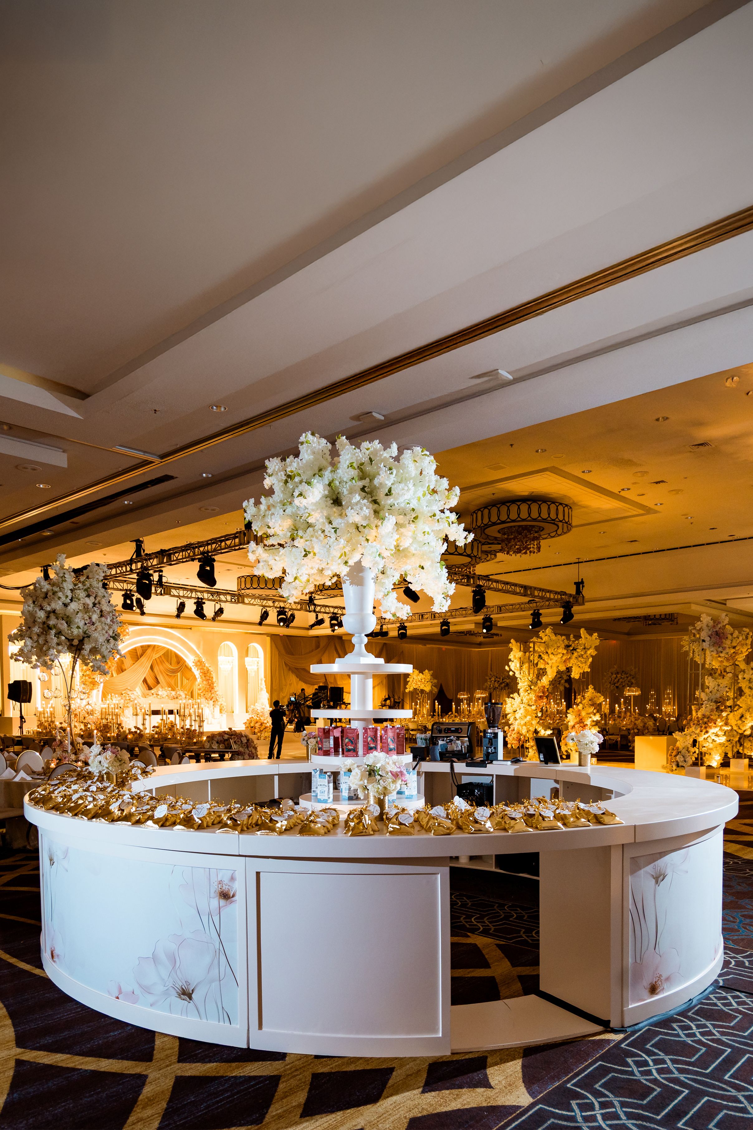Elegant white circular dessert display table with floral arrangements and golden lighting at a luxury wedding reception venue.