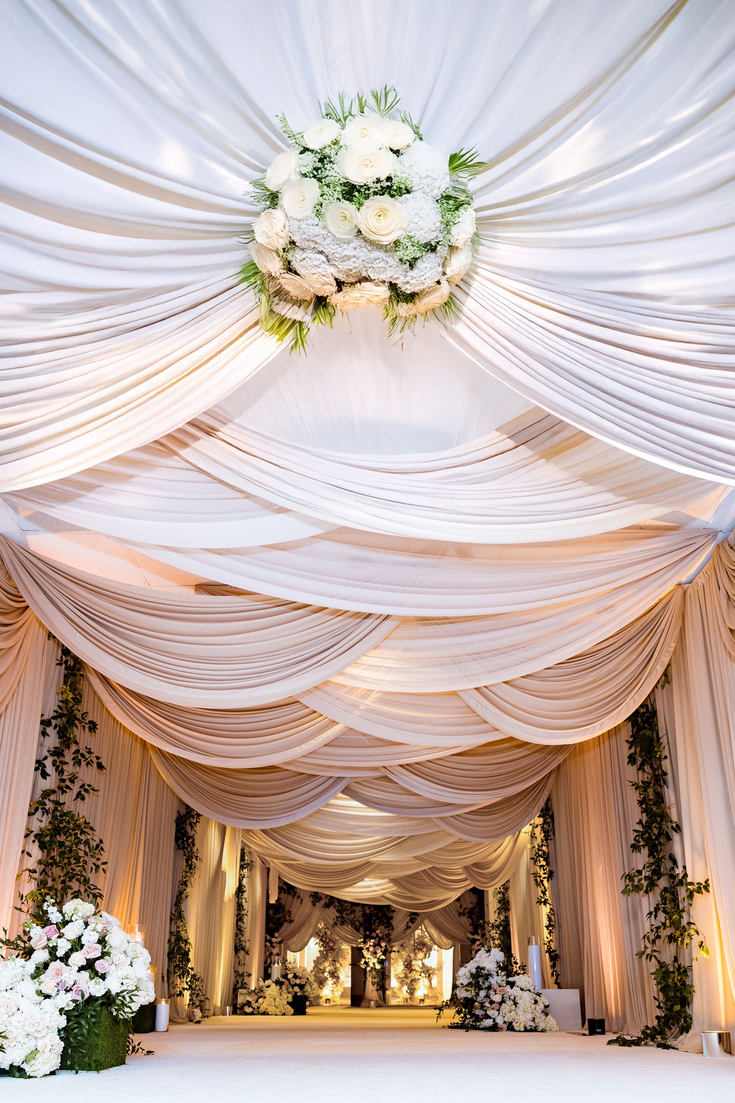 Elegant white fabric draping creates a luxurious ceiling canopy for an indoor wedding reception space with floral accents.