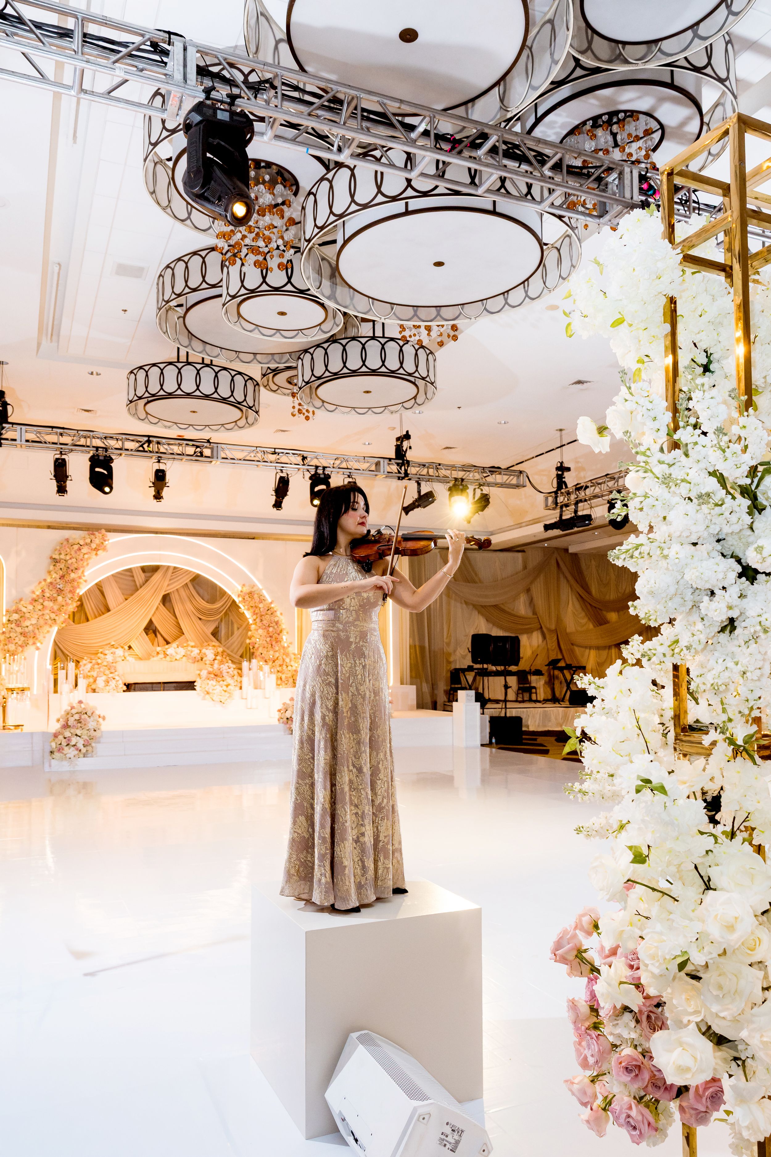 Elegant ballroom with ornate ceiling medallions, white floral arrangements, and a figure in a sparkly gown on a pedestal.