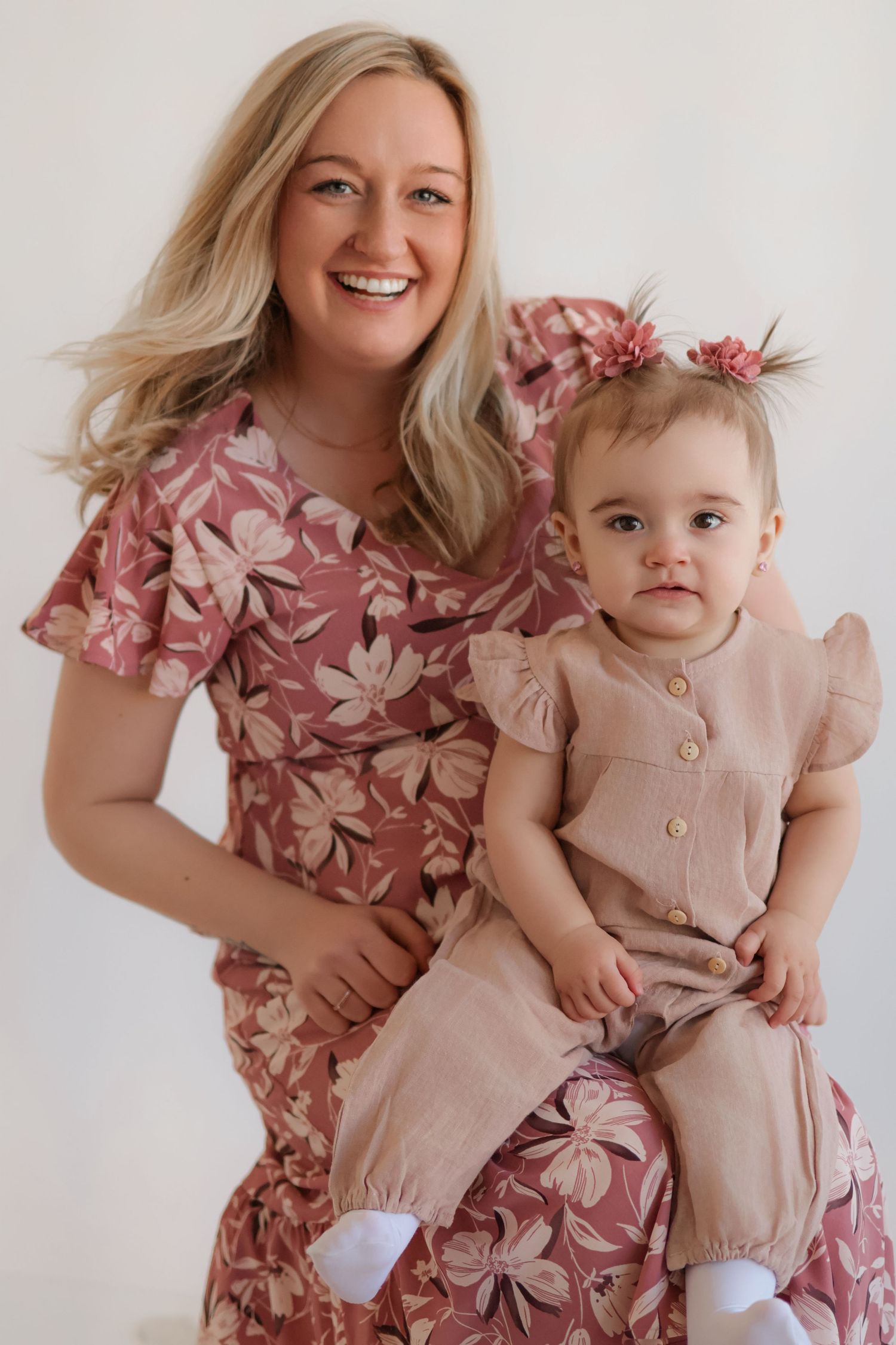 Mother and baby in coordinating pink floral dresses posing together against white backdrop.
