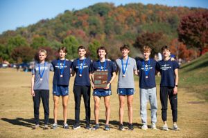 Cross country team members wearing blue uniforms pose with medals and a trophy on a sunny autumn field.
