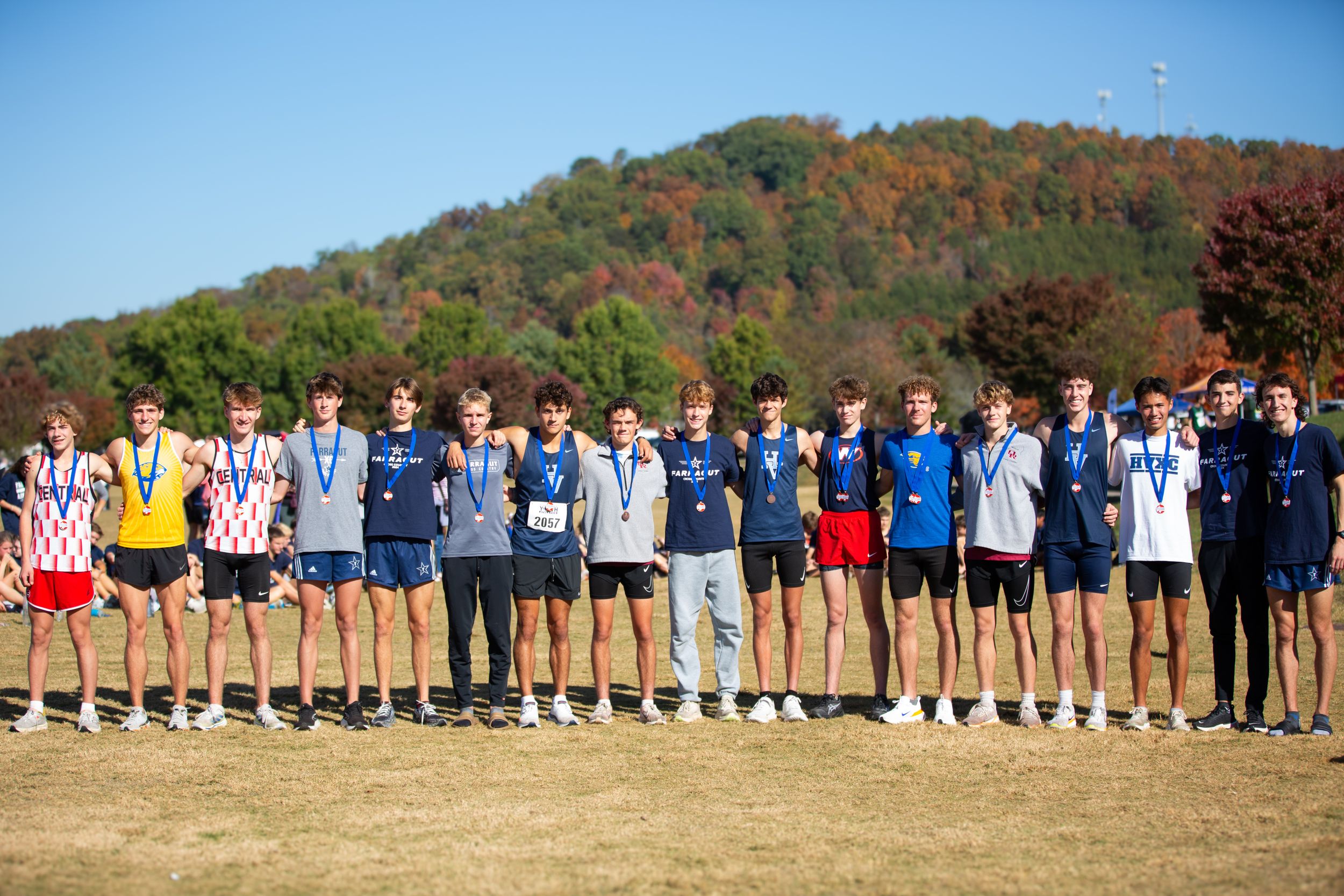 Cross-country team stands in line outdoors against backdrop of autumn foliage on hillside.
