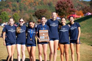 Group of teammates wearing matching navy blue uniforms pose together on sunny grassy hill with autumn trees in background.