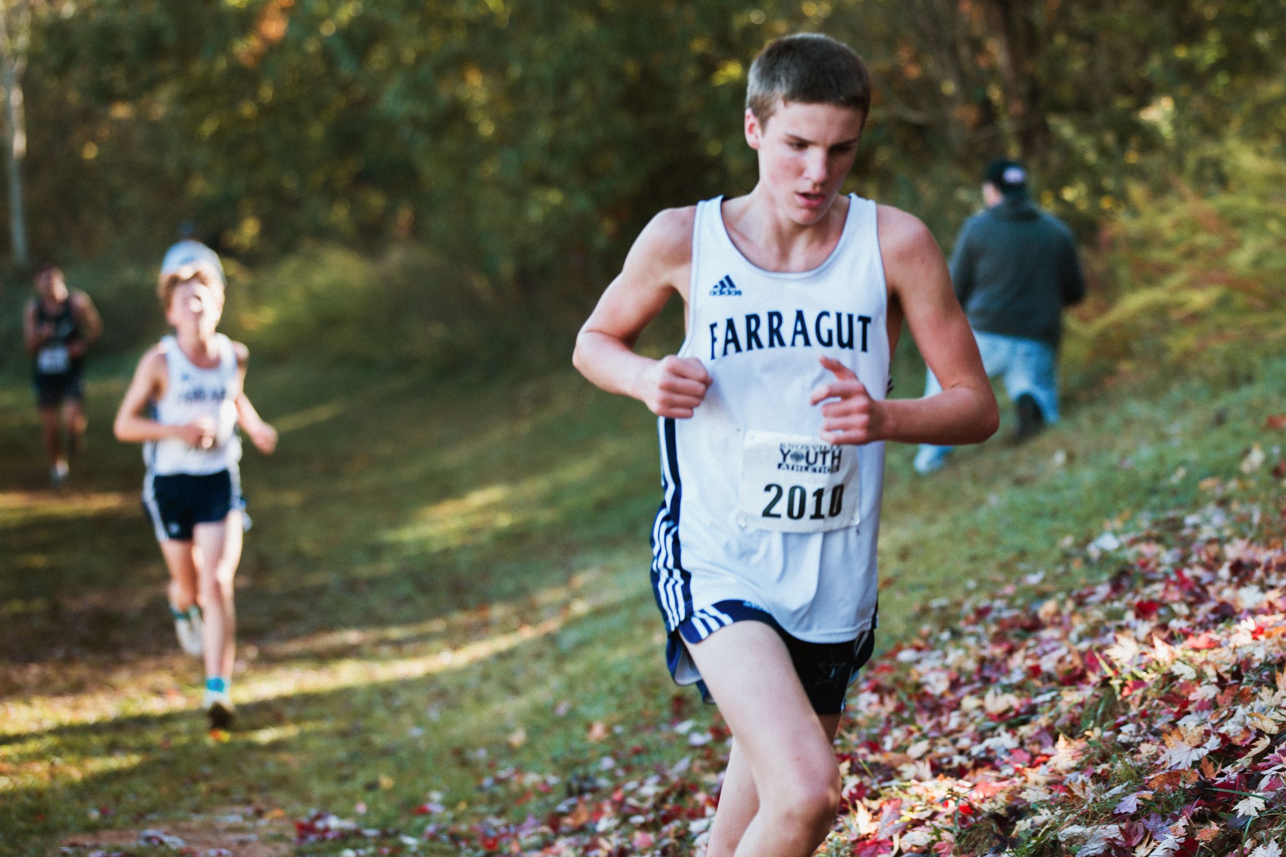 Cross country runner in white Barragh jersey competes on wooded fall trail with another athlete visible in background.