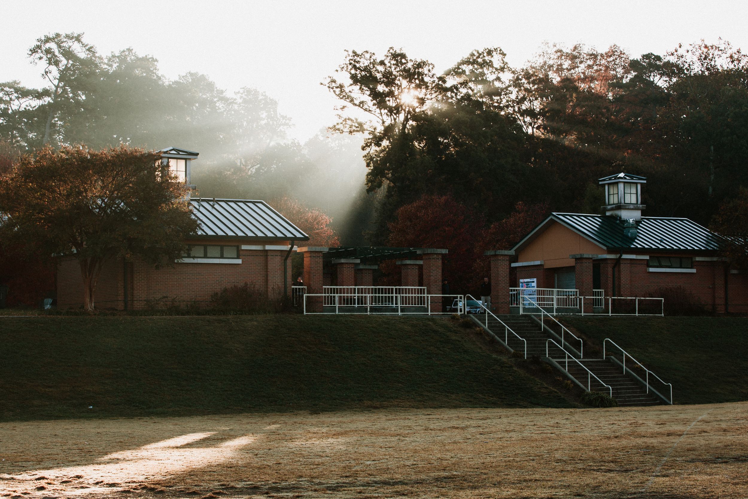 Morning sunbeams pierce through trees onto rustic buildings with metal roofs and white railings at sunset.