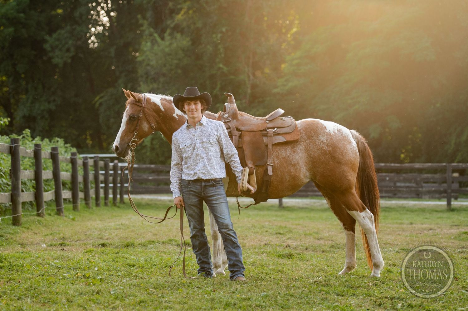 A person stands with a brown and white paint horse on a fenced pasture at sunset.