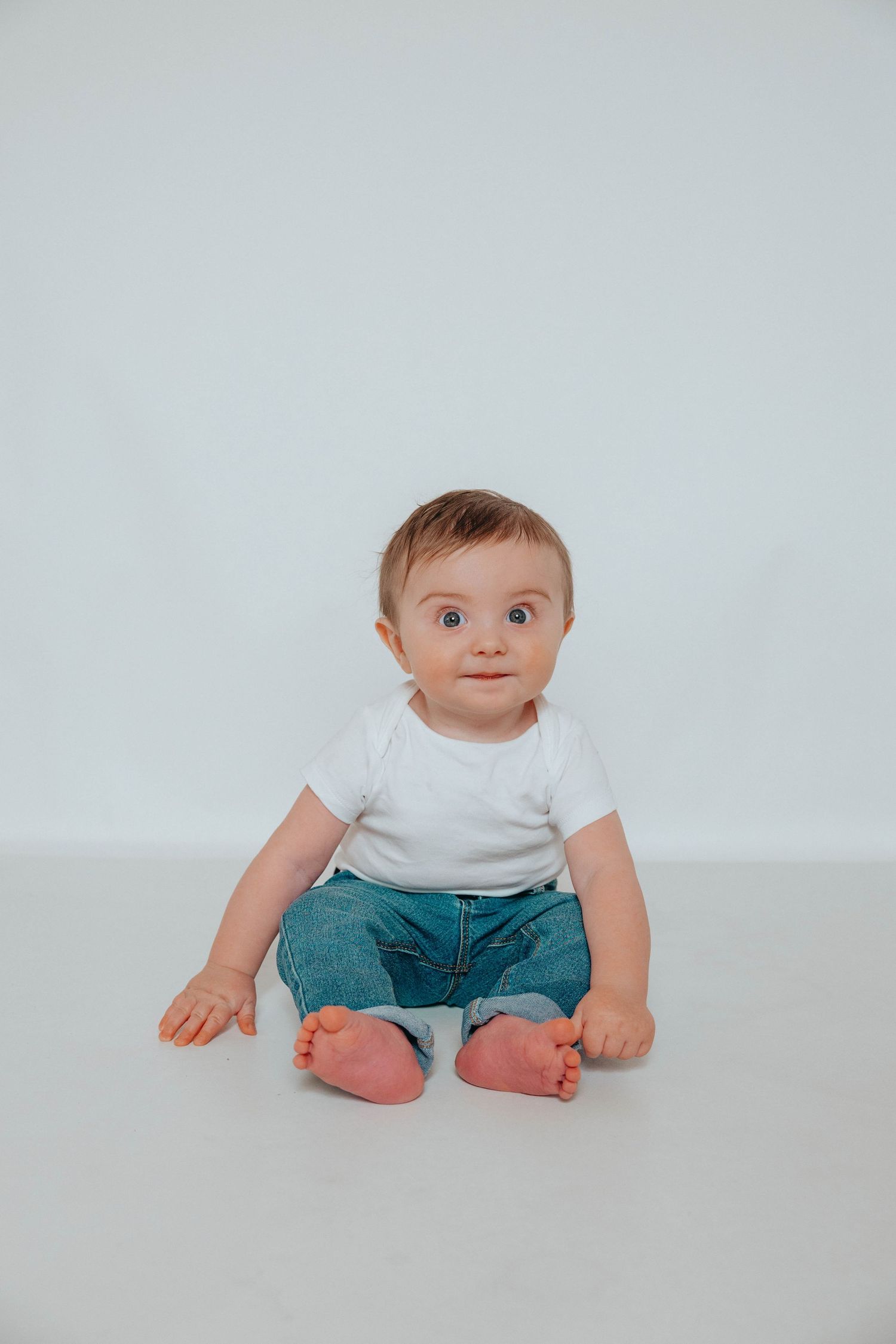Baby wearing white shirt and teal shorts sitting on light background with eager expression.