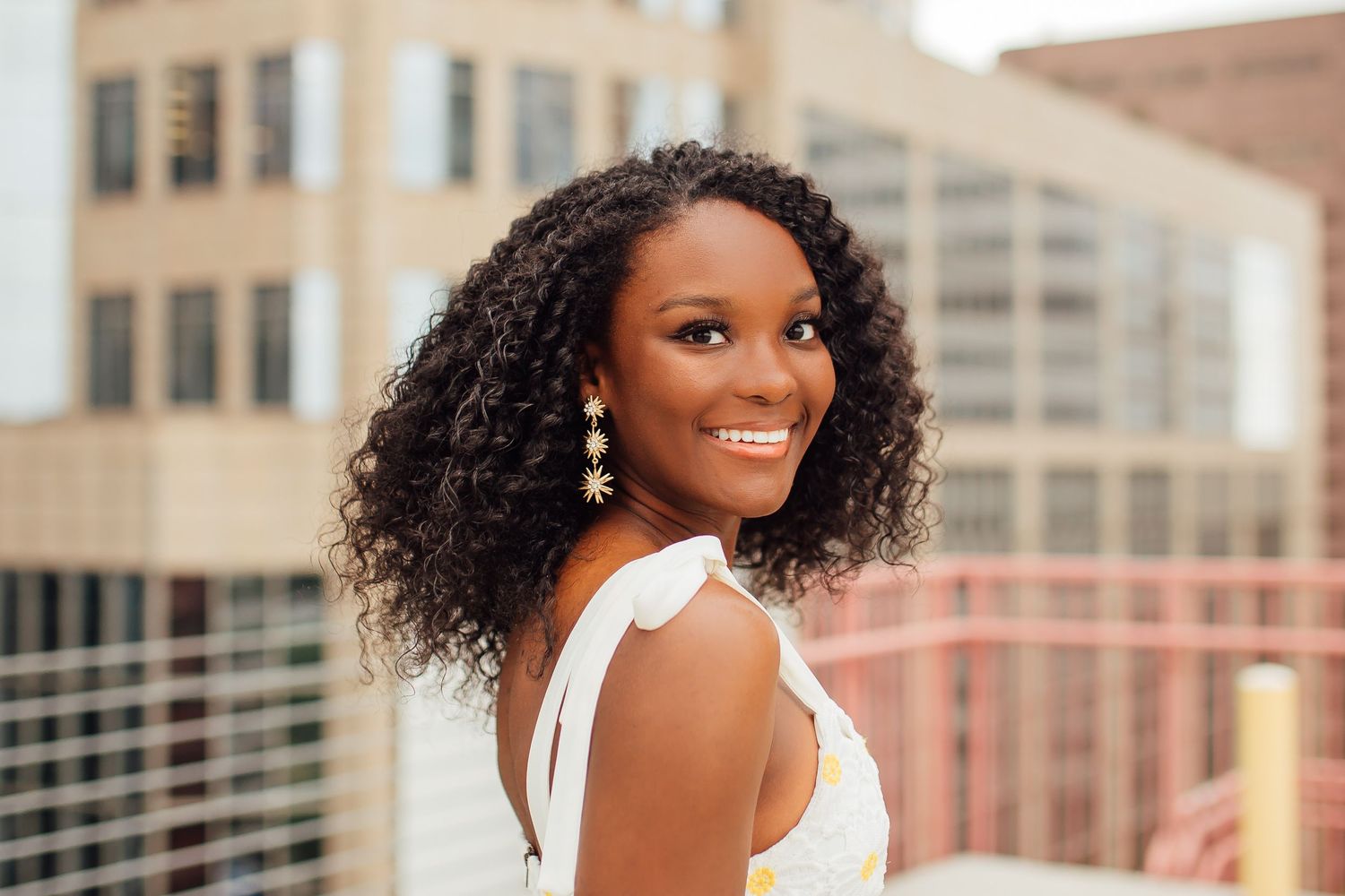 A smiling woman with curly natural hair poses outdoors in a white sleeveless top against an urban backdrop.