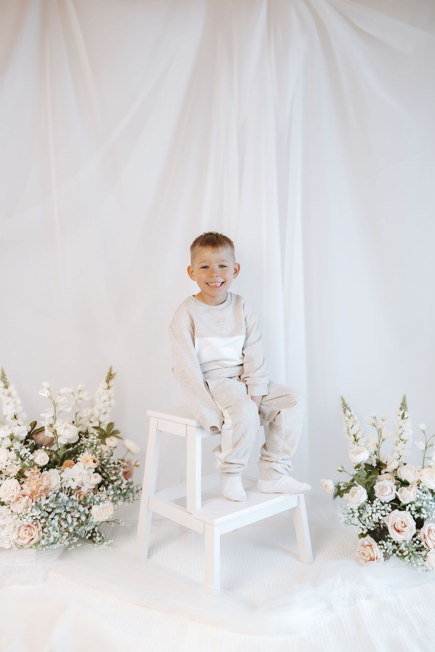 A white decorated studio setting with floral arrangements and a small white stepstool against a light backdrop.