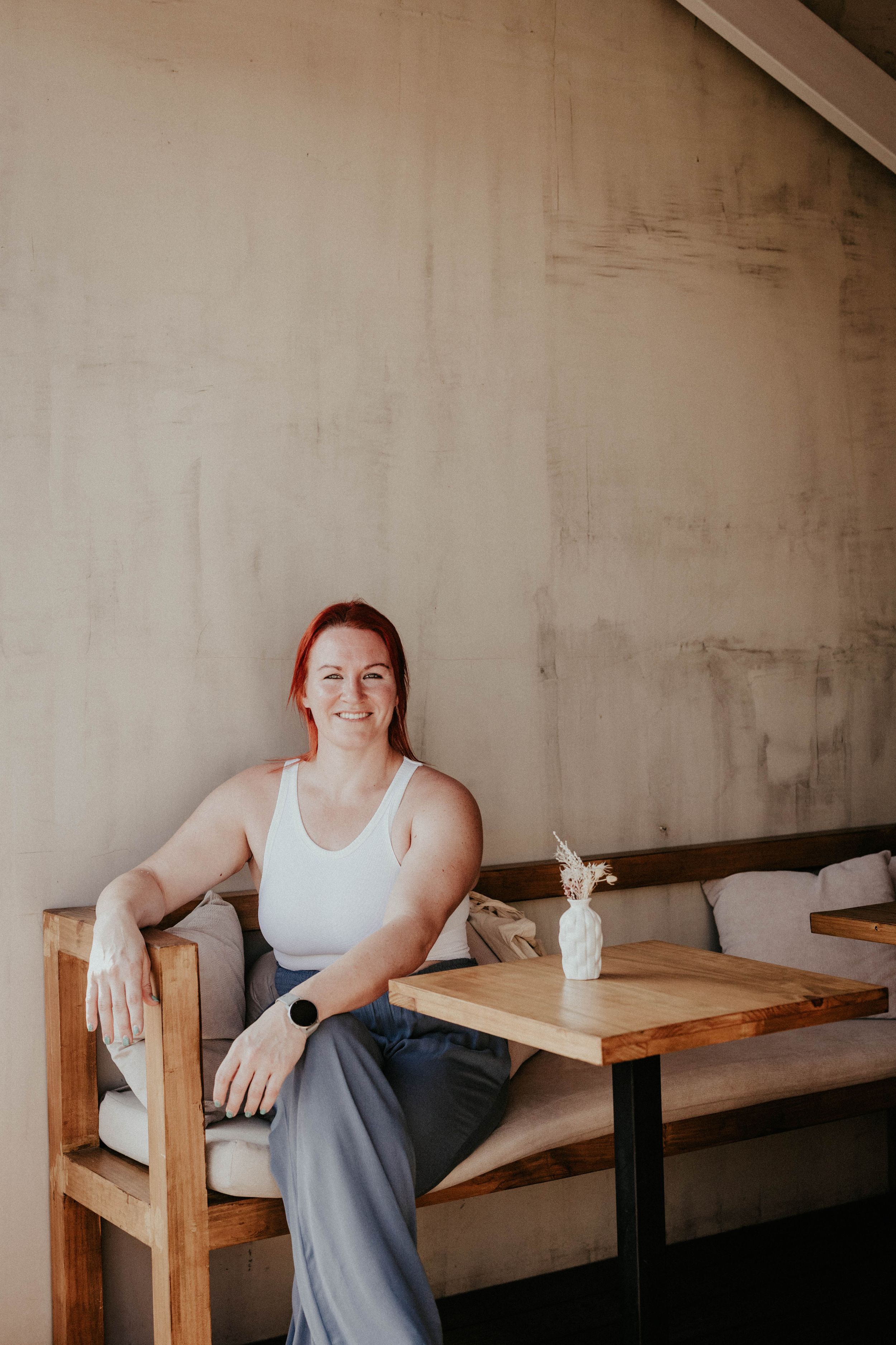 Person wearing white tank top sits on rustic wooden bench against concrete wall in minimalist interior setting.